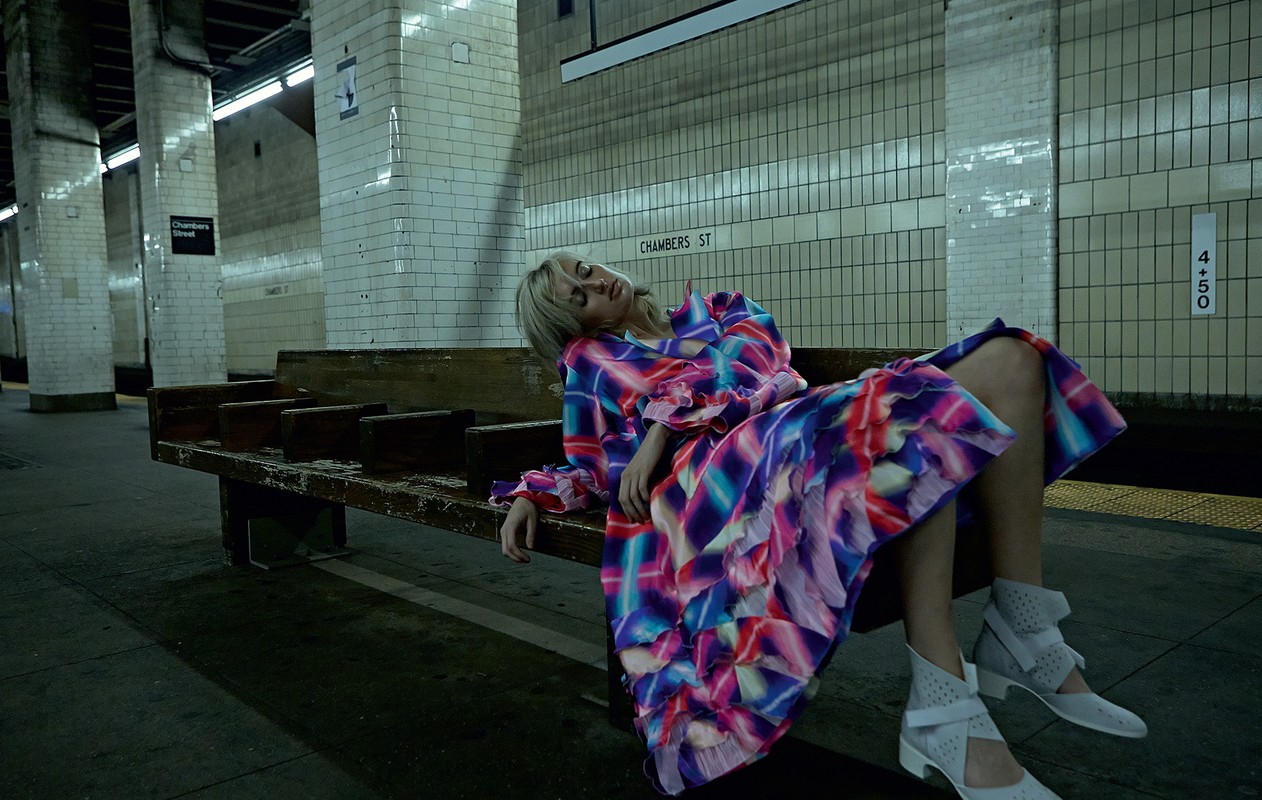 Woman in colourful dress reclines on subway bench in urban setting. The underground platform features tiled walls and dim lighting.