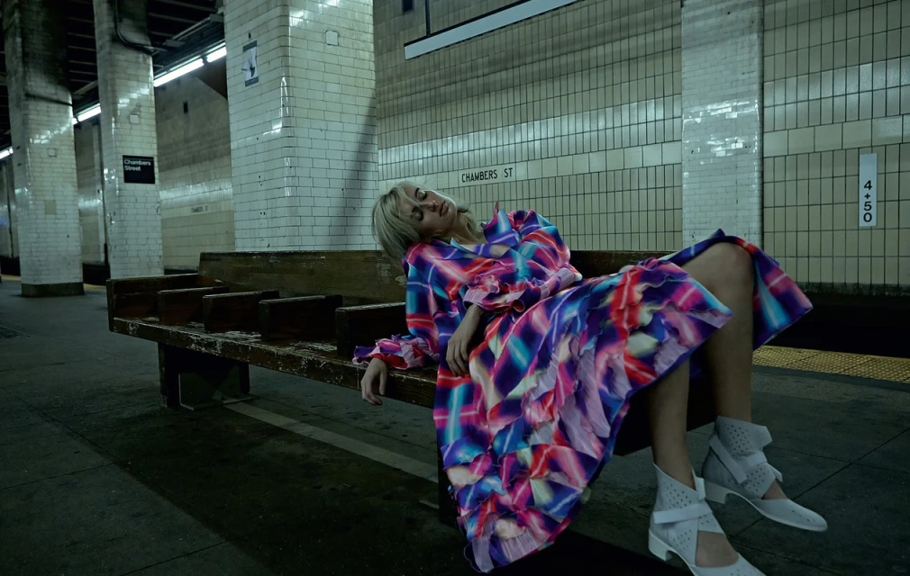 Woman in colourful dress reclines on subway bench in urban setting. The underground platform features tiled walls and dim lighting.