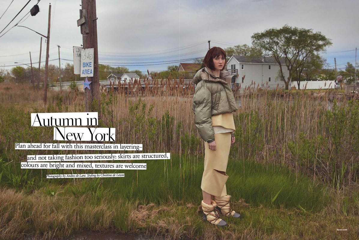 Fashion editorial shot outdoors: adult woman poses wearing a cropped puffer jacket, skirt with boots, tall reeds in background.