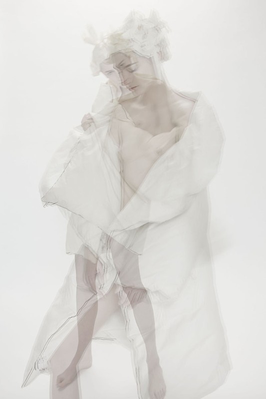 Artistic ethereal photo of a woman in a sheer white robe and floral headpiece against a soft white background with a soft, dreamy aesthetic.