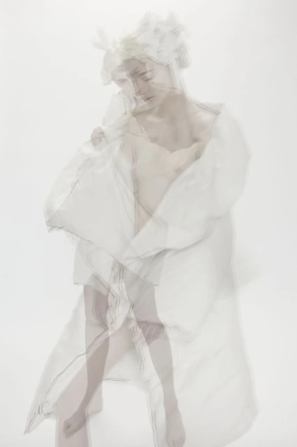 Artistic ethereal photo of a woman in a sheer white robe and floral headpiece against a soft white background with a soft, dreamy aesthetic.