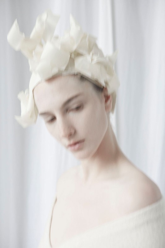 Close-up of an adult female model wearing an off-white headpiece and shawl against an off-white curtain backdrop, gazing down.