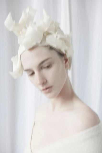 Close-up of an adult female model wearing an off-white headpiece and shawl against an off-white curtain backdrop, gazing down.