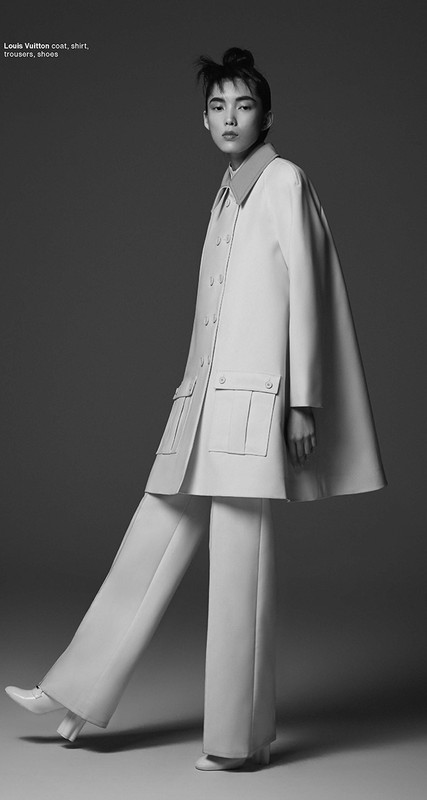 Model in a structured light-colored coat and wide trousers standing against a gradient studio background.