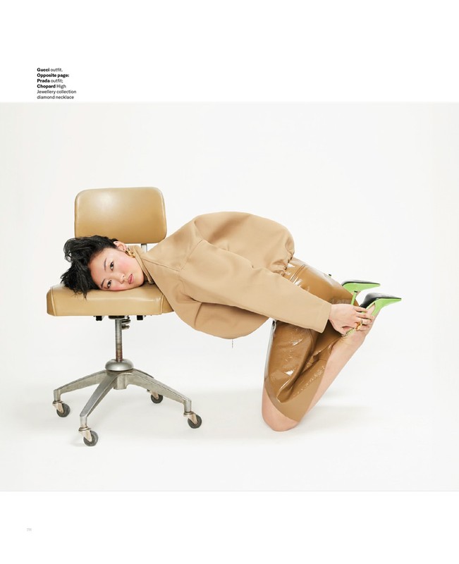 Woman in tan jacket and shiny pants poses on tan chair in a white studio. She is kneeling on the floor with her face on the chair.