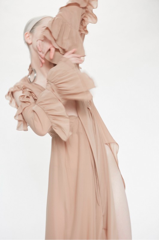 Woman in a peach-colored ruffled dress with long sleeves stands against a bright white background in a fashion or editorial shot.