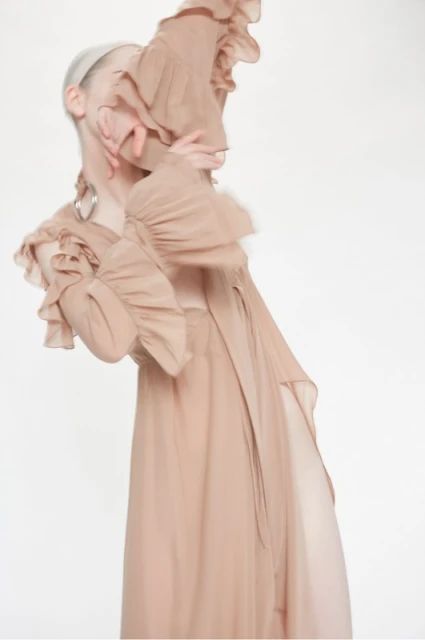 Woman in a peach-colored ruffled dress with long sleeves stands against a bright white background in a fashion or editorial shot.