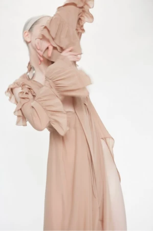 Woman in a peach-colored ruffled dress with long sleeves stands against a bright white background in a fashion or editorial shot.