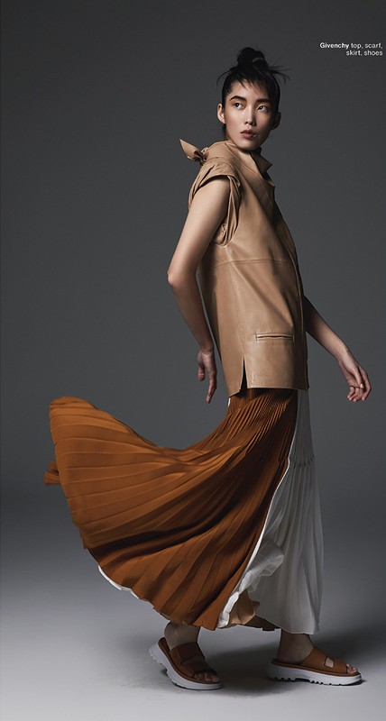 A stylish Asian woman poses in a tan sleeveless top and pleated skirt in a studio, showcasing a contemporary fashion look.