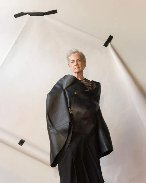 Older woman with short gray hair poses in a black top and layered leather wrap in front of a white backdrop with black tape.