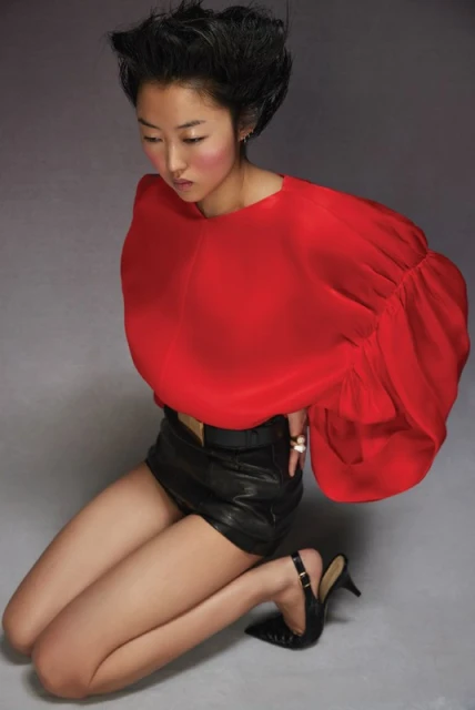 A woman with dark hair kneels in a photo studio. She wears a red blouse with large sleeves and black shorts, styled with high-heel shoes.