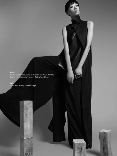Fashion shot of an Asian woman in a black sleeveless coat and wide-leg pants, posing near stacked wooden blocks against a grey background.