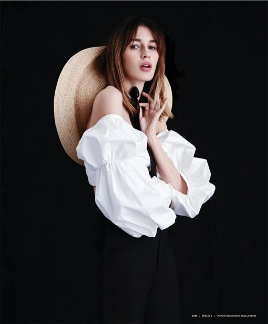 A woman wearing a white peasant blouse, black pants, and a straw hat poses against a dark backdrop in a fashion editorial shot.