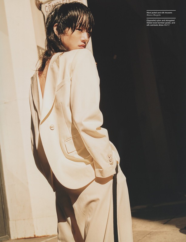 Elegant Asian woman poses in an off-white suit near a dark doorway, bathed in warm light. The suit features a relaxed fit with wide-leg pants.