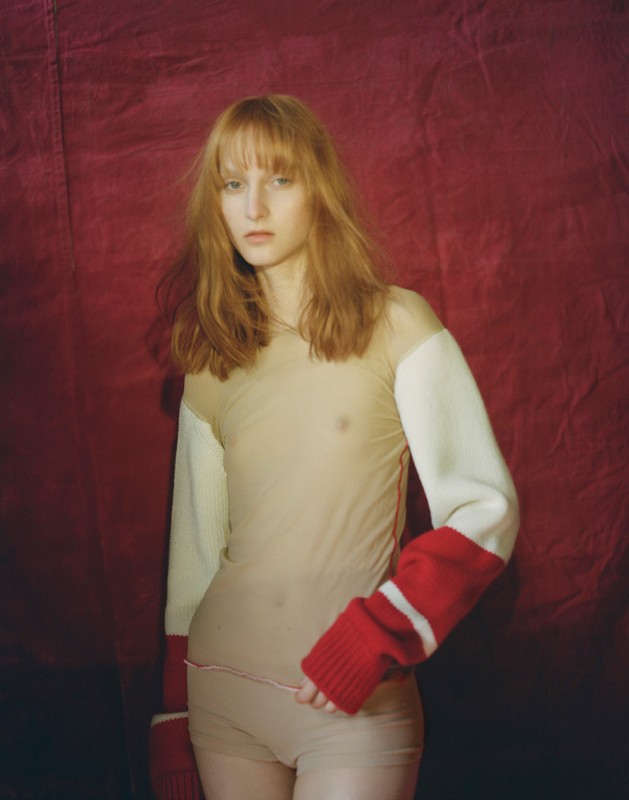 Woman with red hair poses in neutral briefs and a sheer top with red and white knitted sleeves in front of a red backdrop.