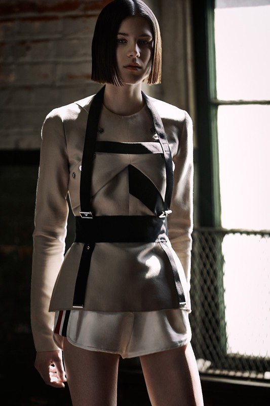 A fashion shot of an adult female model wearing a beige jacket with black harness, standing in front of a brick wall and window.