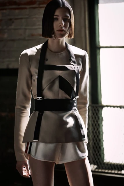 A fashion shot of an adult female model wearing a beige jacket with black harness, standing in front of a brick wall and window.