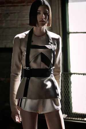 A fashion shot of an adult female model wearing a beige jacket with black harness, standing in front of a brick wall and window.