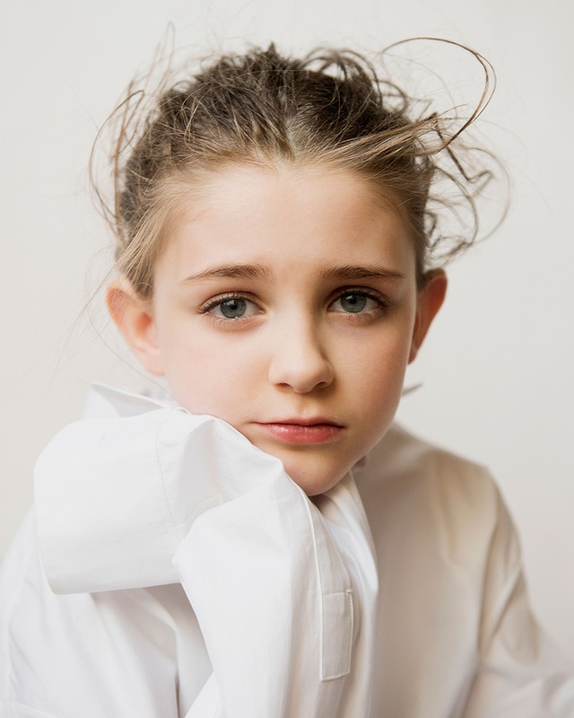 Portrait of a young girl with messy hair wearing a white shirt looking directly at the camera against a plain neutral background.