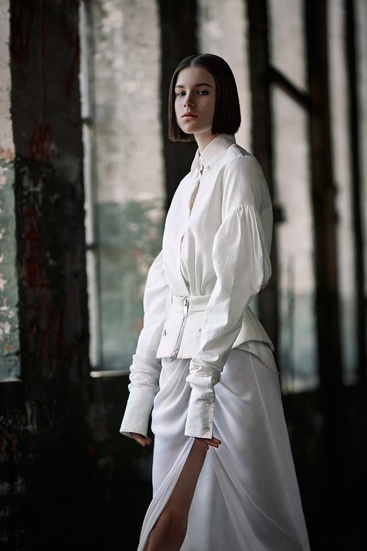 Woman with short dark hair wearing a white blouse and skirt against a rustic columned interior background