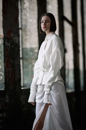 Woman with short dark hair wearing a white blouse and skirt against a rustic columned interior background