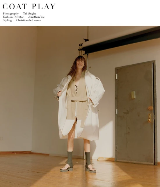 An adult woman poses in a photo studio wearing a beige outfit and a white trench coat, accessorized with socks and sandals on a sunny day.