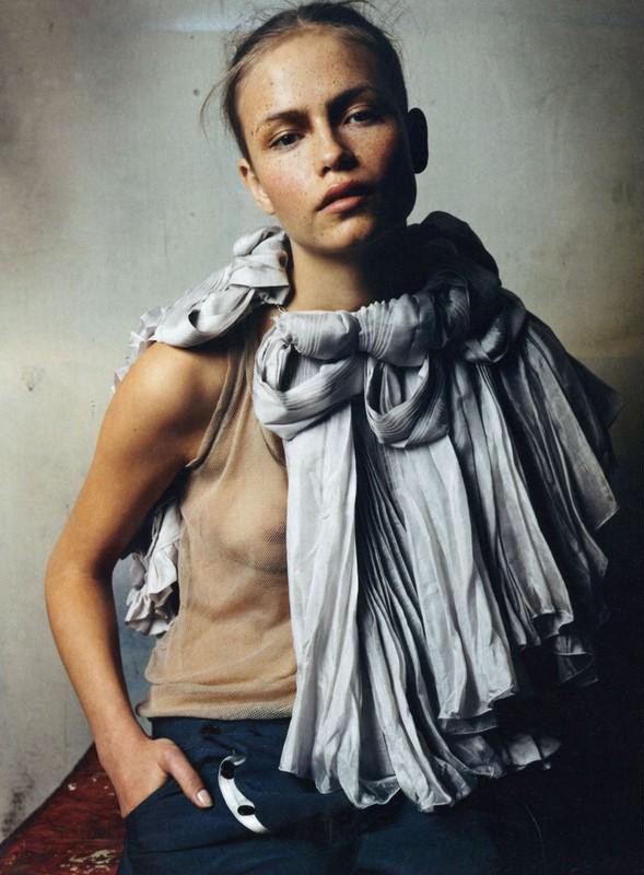 A fair-skinned young woman poses indoors wearing a beige top and a large grey scarf draped around her neck and shoulders.
