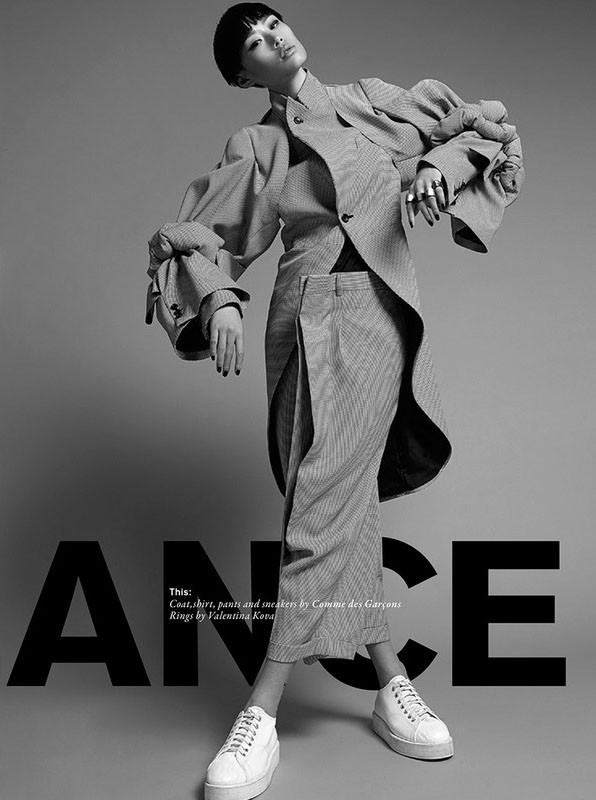 Editorial fashion shot of an Asian female model wearing a grey pinstripe suit with exaggerated sleeves and white platform sneakers indoors.