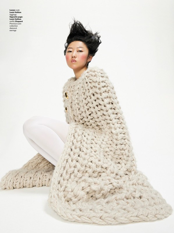 Editorial fashion shot of a seated Asian woman wearing a beige, chunky-knit sweater dress with white tights against a white background.