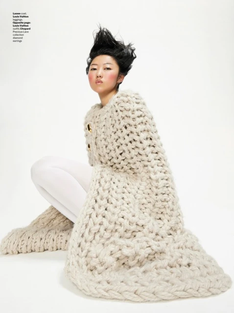 Editorial fashion shot of a seated Asian woman wearing a beige, chunky-knit sweater dress with white tights against a white background.