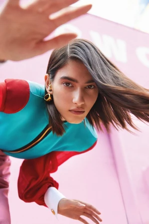 Close-up of a model in a turquoise top with red sleeves, wearing large gold hoop earrings, against a pink urban backdrop.