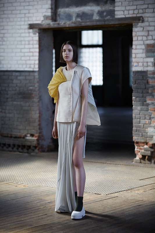 Fashionable woman with short hair poses in a modern sleeveless top and long skirt in front of a brick doorway inside a rustic building.