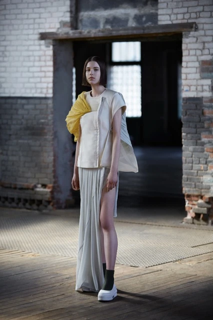 Fashionable woman with short hair poses in a modern sleeveless top and long skirt in front of a brick doorway inside a rustic building.