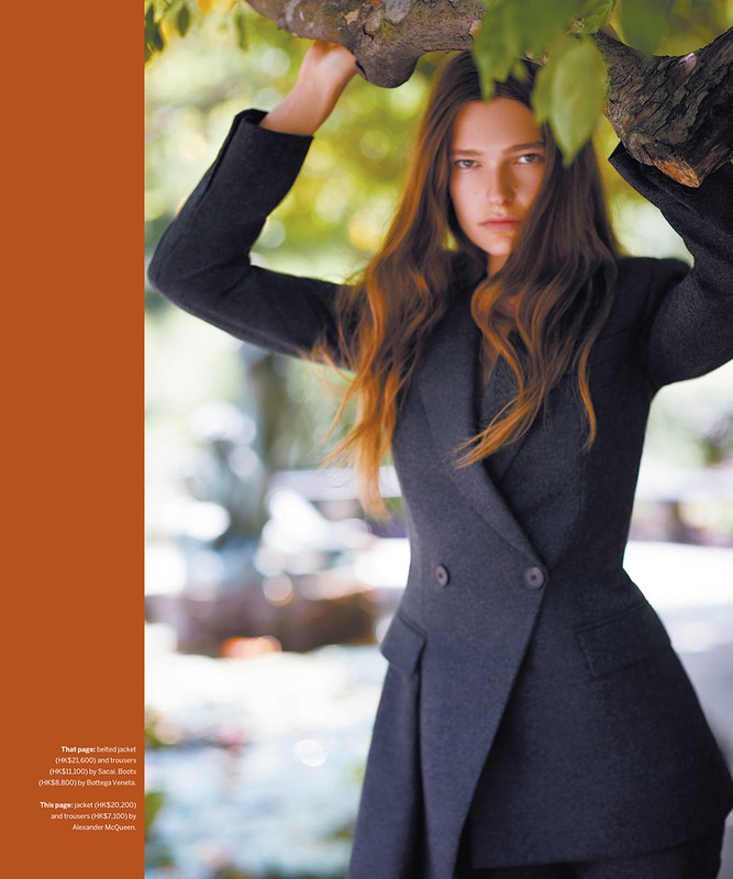 Woman with long brown hair in a gray suit poses beneath a tree branch in an outdoor setting, looking directly at the camera.