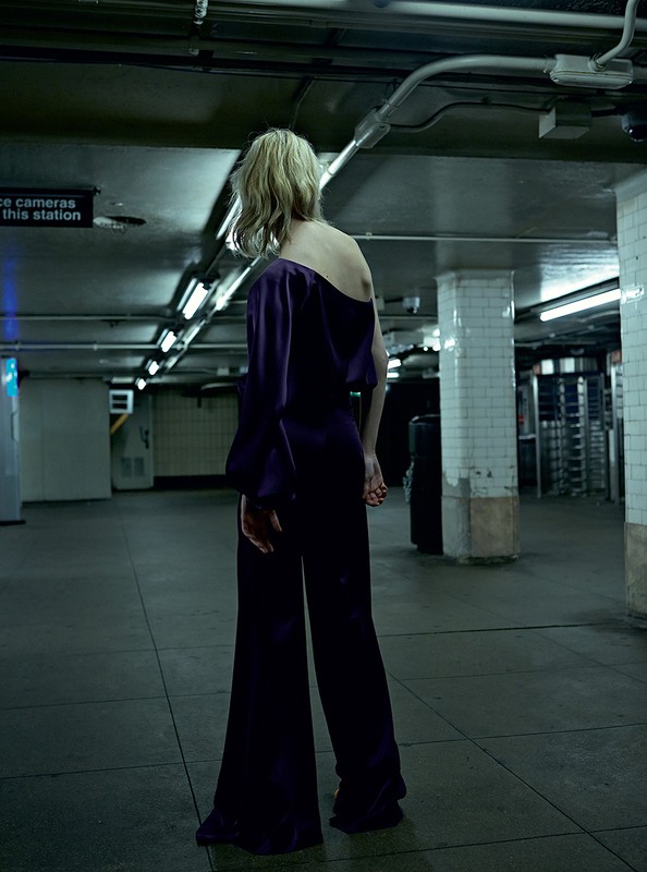 Woman in purple pantsuit in subway station. She faces away from the camera, showcasing the outfit's design and the station's architecture.