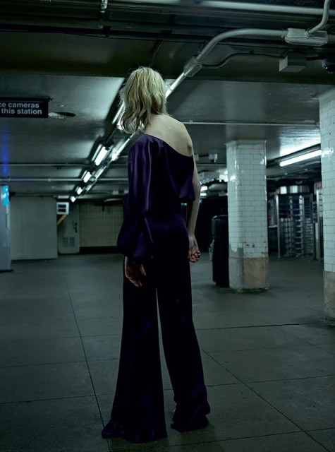 Woman in purple pantsuit in subway station. She faces away from the camera, showcasing the outfit's design and the station's architecture.