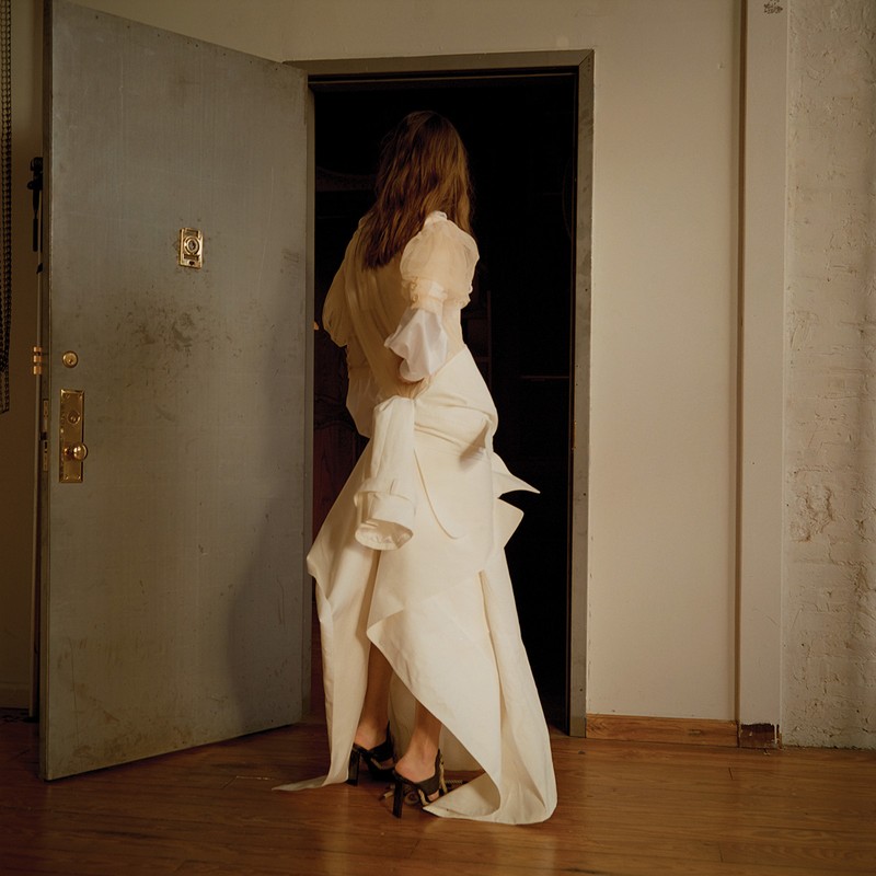 A woman in an avant-garde white dress with a high-low hemline walks through a doorway. The lighting is soft and the setting is indoors.