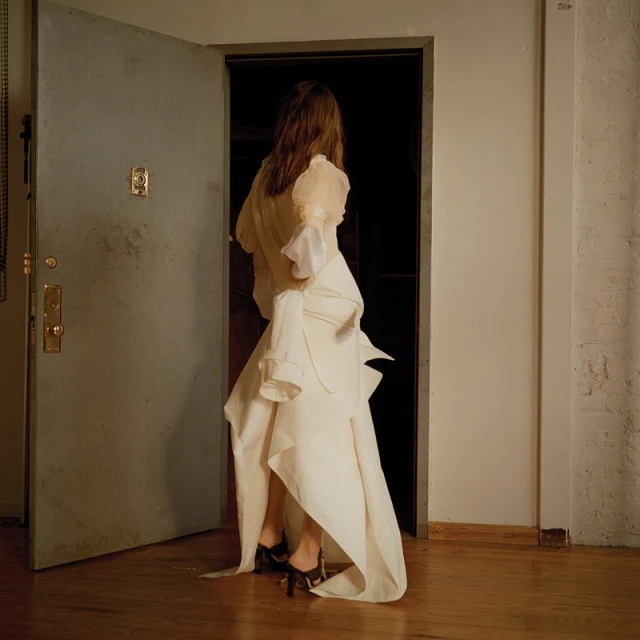A woman in an avant-garde white dress with a high-low hemline walks through a doorway. The lighting is soft and the setting is indoors.