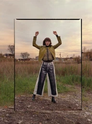 Model in olive jacket and patterned wide leg pants standing outdoors with arms raised