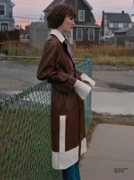 Woman in brown leather trench coat with white cuffs leaning against chain-link fence outdoors with houses.