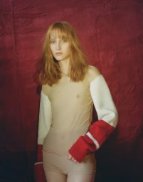 Model with long reddish hair wearing a sheer nude top with white and red sleeves against a dark red backdrop