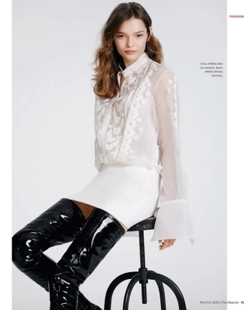 Model seated on stool wearing sheer white lace blouse and shiny black patent leather over-the-knee boots.
