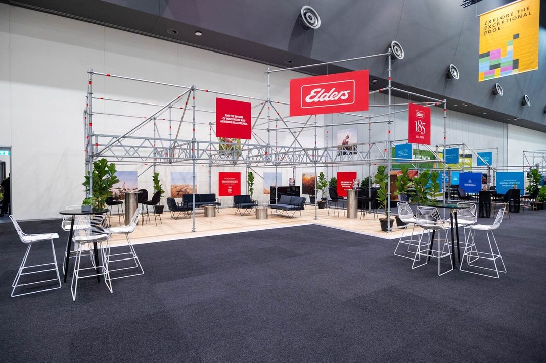 Interior of a convention hall featuring a brand exhibition space with tables, chairs, plants, and brand signage under a metal frame.