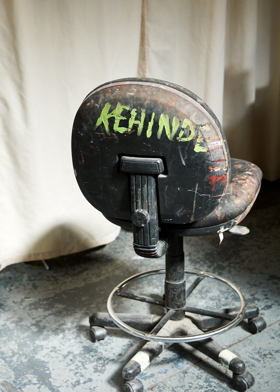 An industrial black swivel chair is heavily worn with graffiti, paint splatters, and scuffs, set against a neutral fabric backdrop.