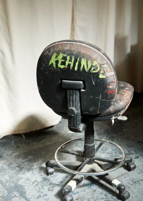 An industrial black swivel chair is heavily worn with graffiti, paint splatters, and scuffs, set against a neutral fabric backdrop.
