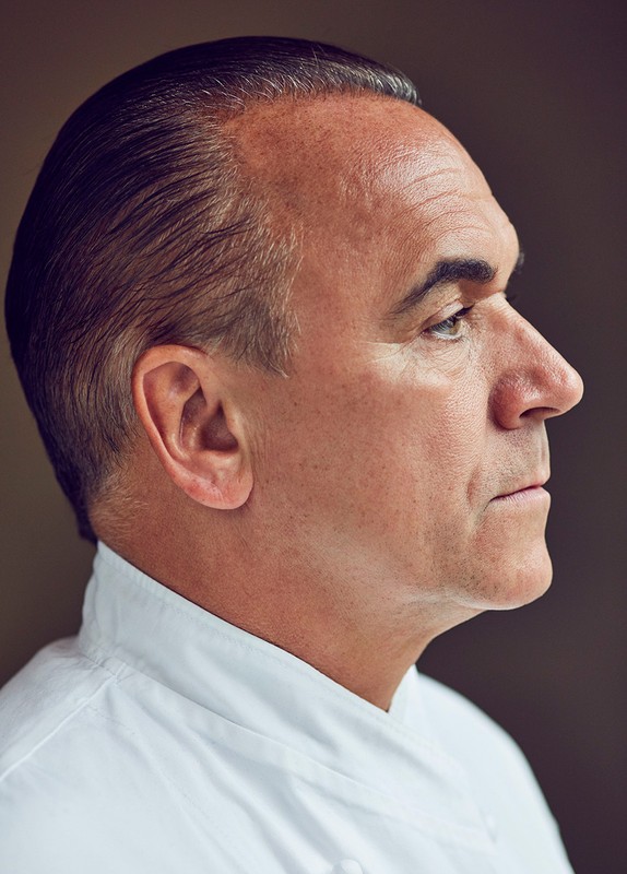 Portrait of a middle-aged man in a white chef's jacket, captured in a side view with soft lighting and a blurred background.
