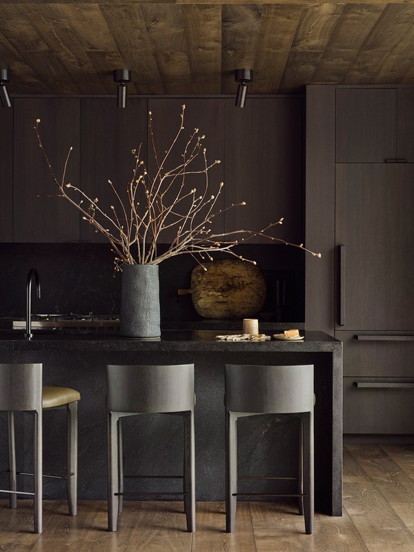 Moody kitchen interior with dark cabinets, wooden ceiling, stone countertop, bar stools, and a decorative vase with branches.