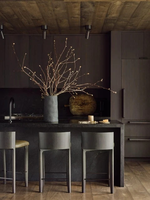 Moody kitchen interior with dark cabinets, wooden ceiling, stone countertop, bar stools, and a decorative vase with branches.