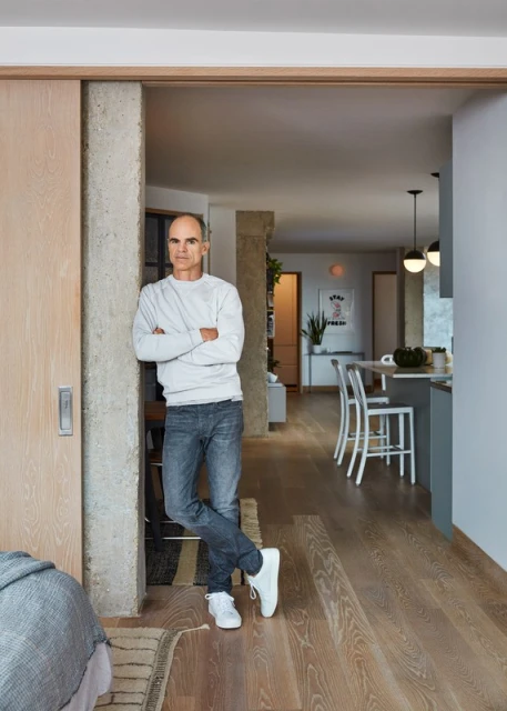 Indoor portrait of a bald adult male in a grey sweatshirt and jeans standing in a modern home, with natural lighting and wooden floors.