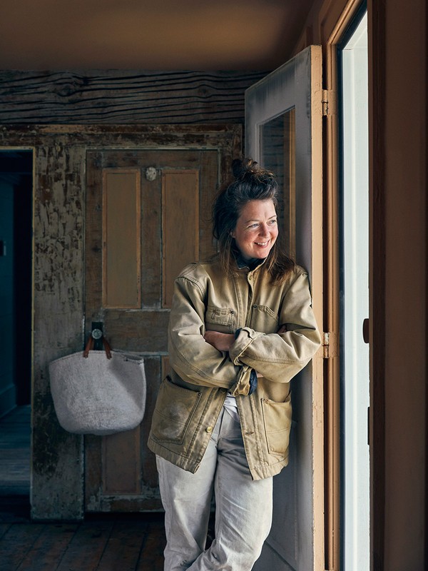 Woman in a tan jacket smiles as she leans against a white-trimmed doorway inside a rustic home with wooden walls and a hanging bag.